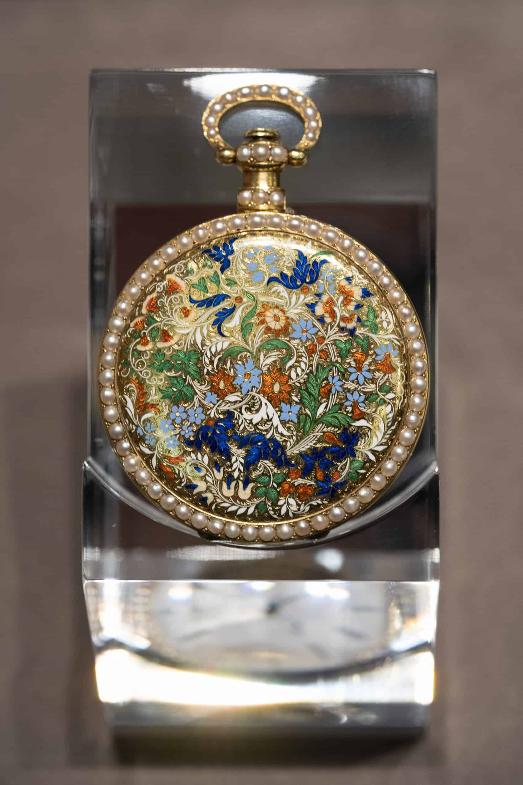 A detailed, ornate pocket timepiece with intricate enamel artwork and a pearl-studded border.