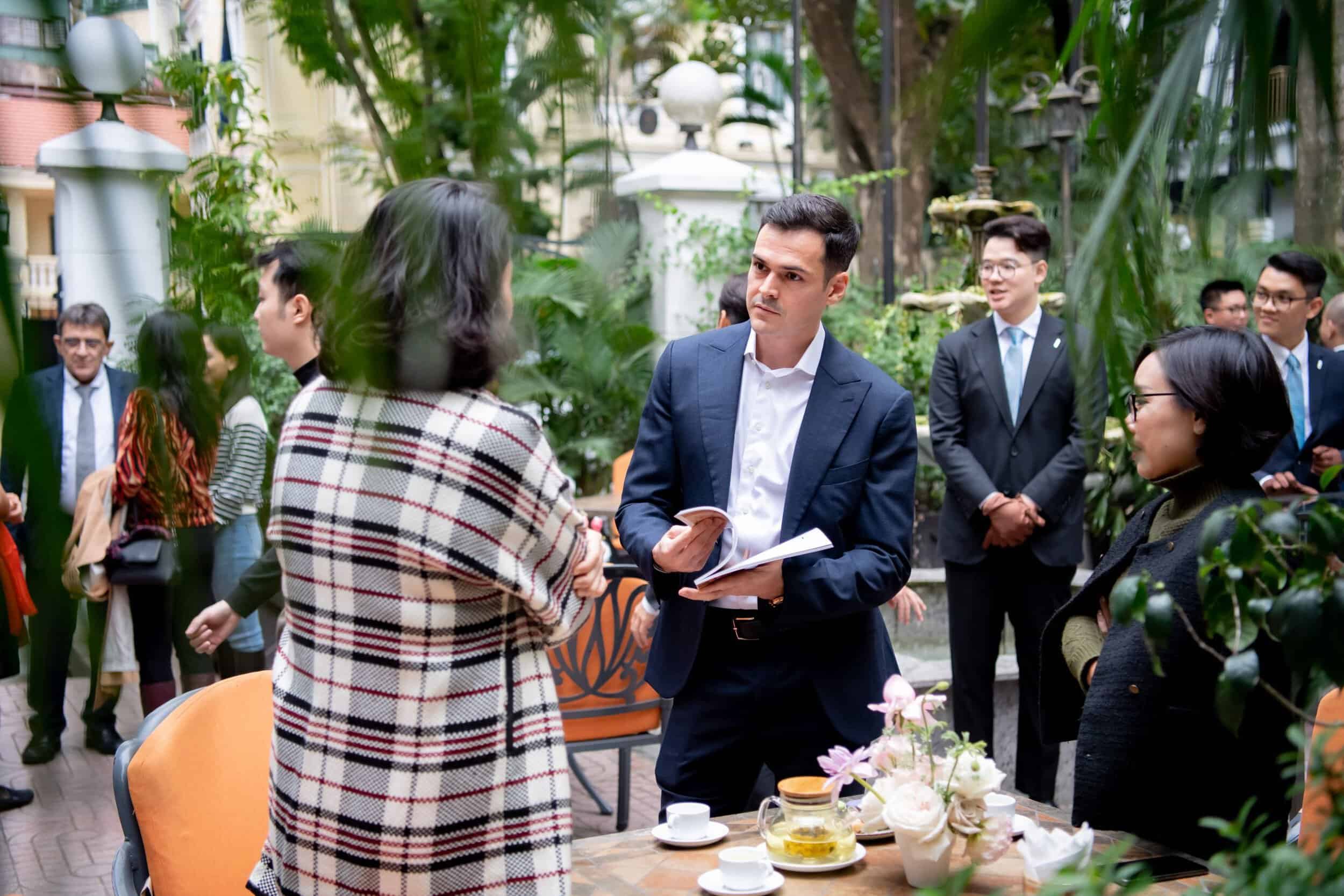 Elegant man discussing with a woman at a social event, surrounded by greenery and other guests, show.